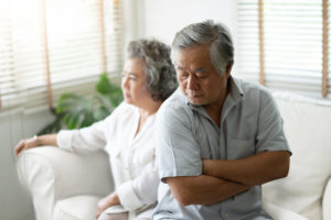 Upset older man sitting on the edge of the sofa with his arms crossed and his wife in the background